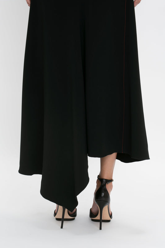 Woman in a Victoria Beckham long black skirt with front-tie detail and high-heeled shoes, standing with one foot slightly behind the other against a white background.