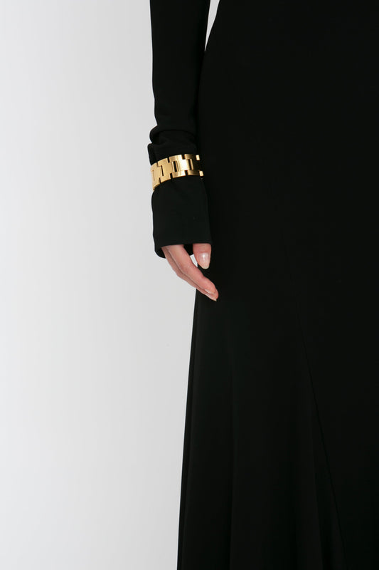 Close-up of a person's arm adorned with a golden chunky bracelet, wearing a black gown with elegant sleeves, against a white background. (Product: Victoria Beckham Tie Detail Floor-Length Dress in Black)