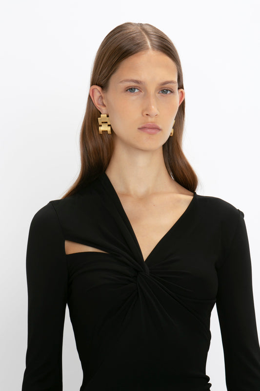 Woman with straight brown hair wearing a Victoria Beckham Tie Detail Floor-Length Dress in Black with asymmetric cut-out and gold earrings, against a white background.