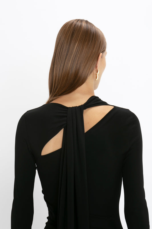 Woman with straight hair, wearing a black Victoria Beckham body-sculpting gown with a back cut-out and a draped detail, seen from behind against a white background.