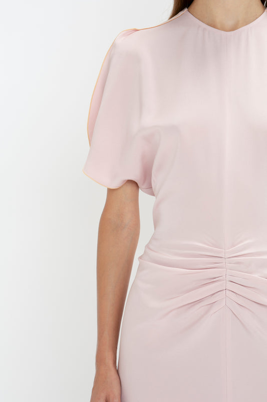 Woman in an elegant Victoria Beckham Gathered Waist Midi Dress In Blush, with tulip sleeves, standing against a white background, cropped to show only her torso and arm.