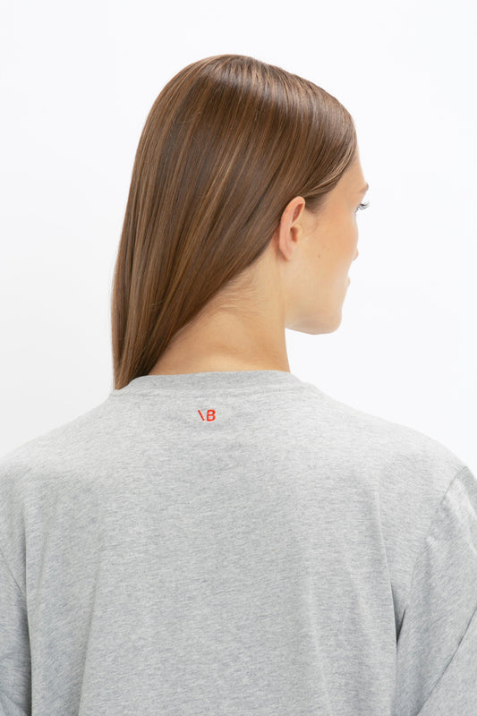 Woman wearing a Asymmetric Relaxed Fit T-Shirt In Grey Marl made of organic cotton, with a small red 'vb' logo on the back of the collar, facing away from the camera by Victoria Beckham.