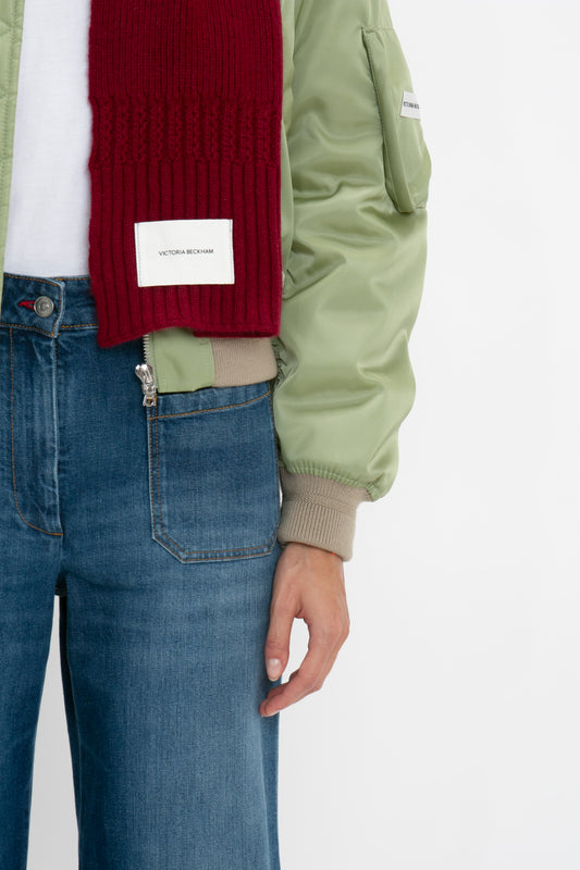 Close-up of a person wearing a green bomber jacket, red sweater, and blue jeans, focusing on their midsection with Victoria Beckham Exclusive Logo Patch Scarf In Burgundy visible.