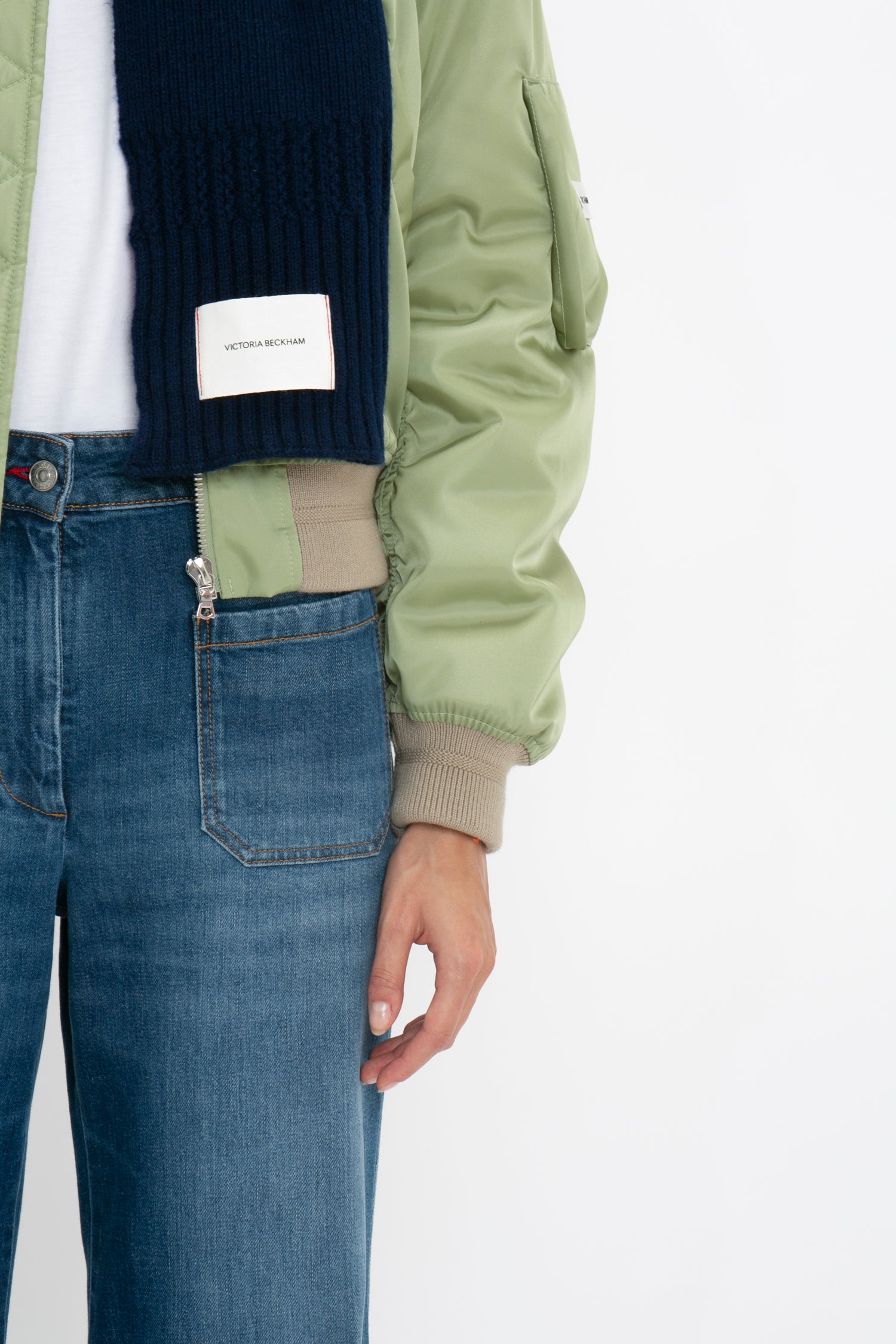 Close-up of a person wearing a green bomber jacket, blue jeans, and a blue sweater with an Exclusive Logo Patch Scarf In Navy by Victoria Beckham visible.