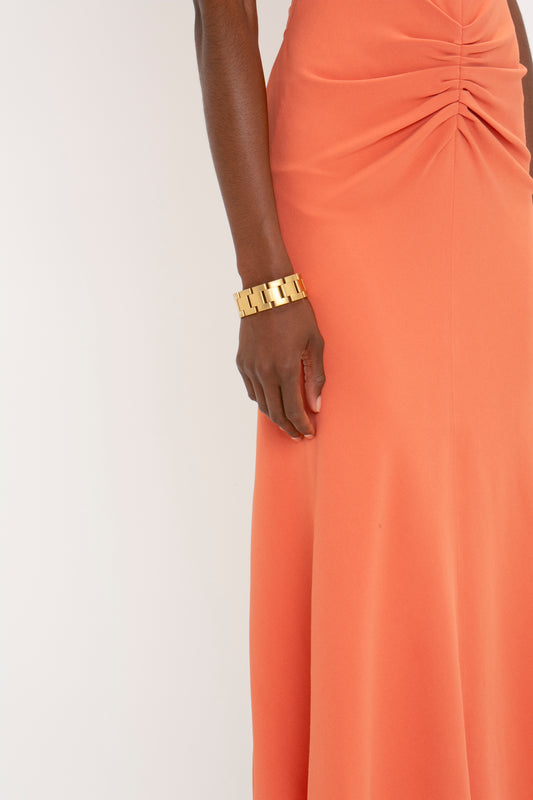 Close-up of a woman's side, wearing a Victoria Beckham Gathered Waist Midi Dress in Papaya and a gold bracelet, focusing on her hand resting elegantly against her hip.