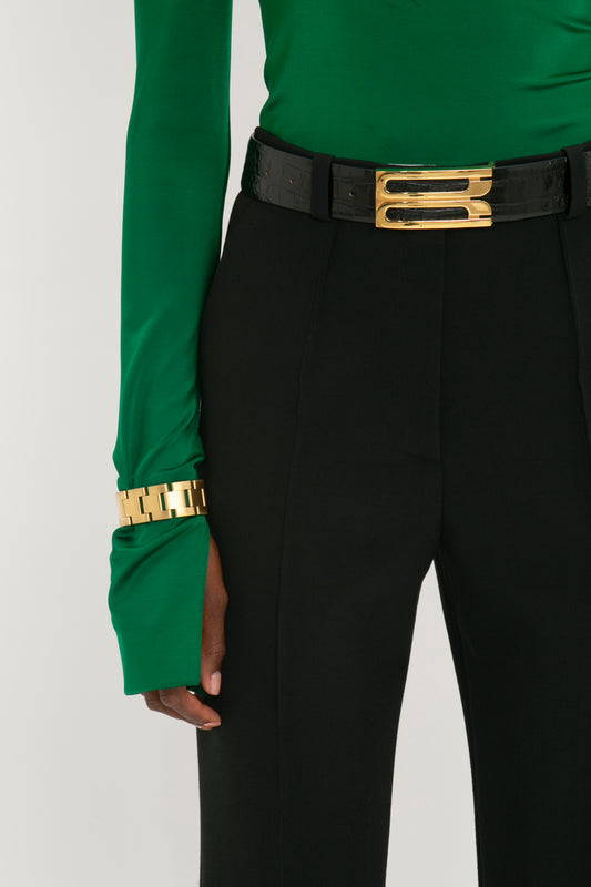Close-up of a woman in a green blouse and black pants, focusing on her Victoria Beckham Jumbo Frame Belt In Black Croc-Effect Leather and matching bracelet.