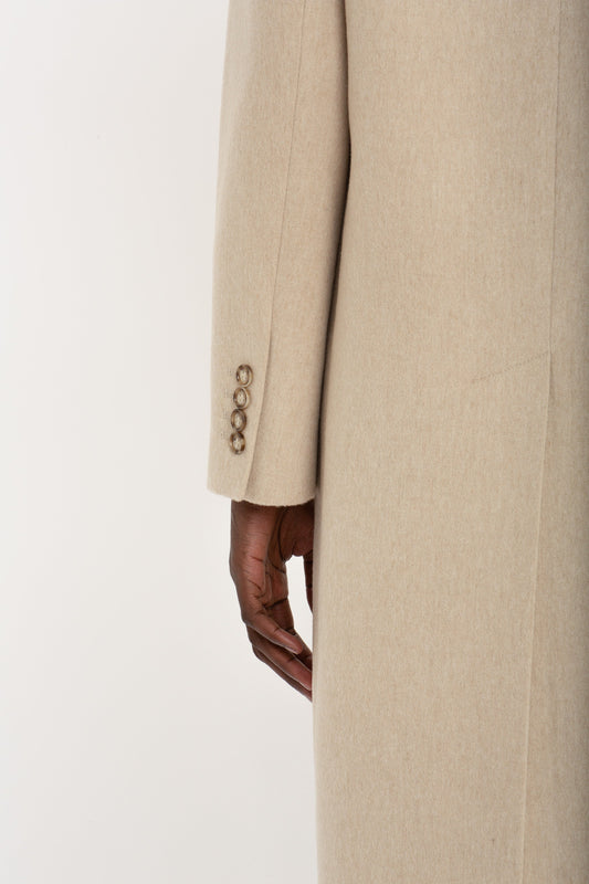 Close-up of a person's side with their hand in the pocket of a beige, wool-cashmere blend Victoria Beckham coat, showing details like buttons and fabric texture.