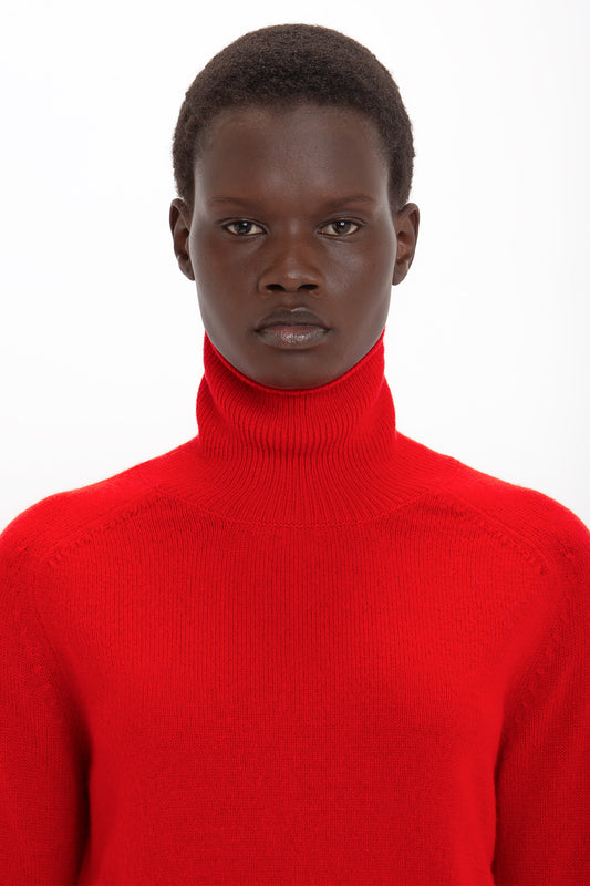 A portrait of a person with short black hair wearing a Victoria Beckham red lambswool polo neck sweater, looking directly at the camera against a white background.