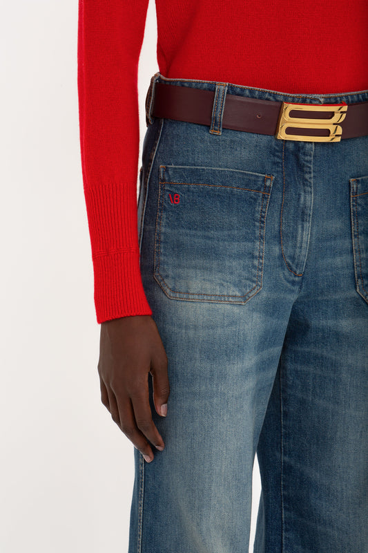 Close-up of a person wearing blue jeans and a Victoria Beckham Luxury Knitwear Red Polo Neck Jumper, focusing on the belt and a hand resting on the thigh.