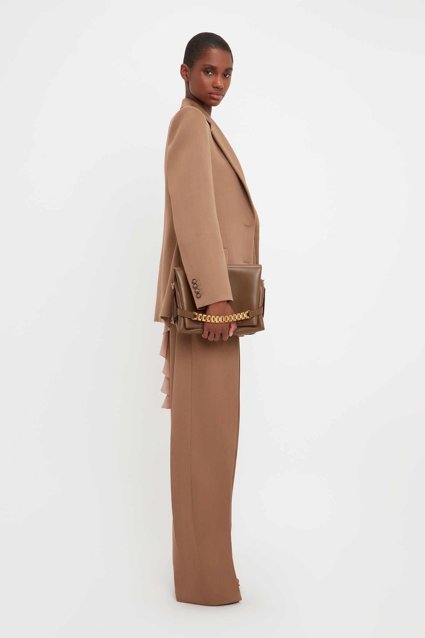 Person wearing a Victoria Beckham Asymmetric Double Layer Jacket In Fawn with a matching brown clutch bag, standing against a white background, showcasing a minimal aesthetic.