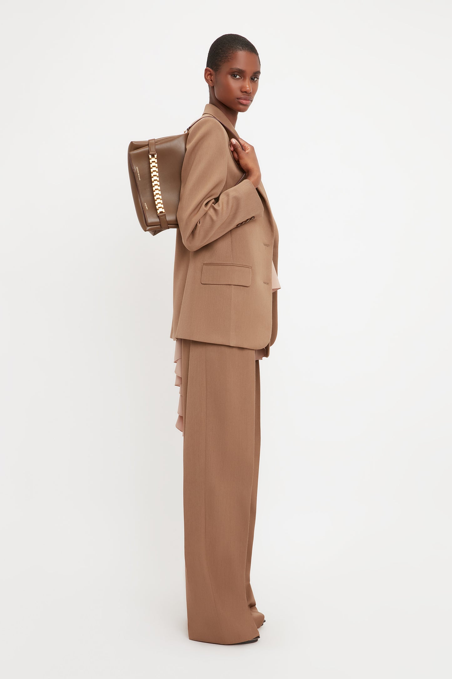 Person in an Asymmetric Double Layer Jacket In Fawn by Victoria Beckham standing sideways, holding a brown handbag featuring a chain detail, showcasing a minimal aesthetic.