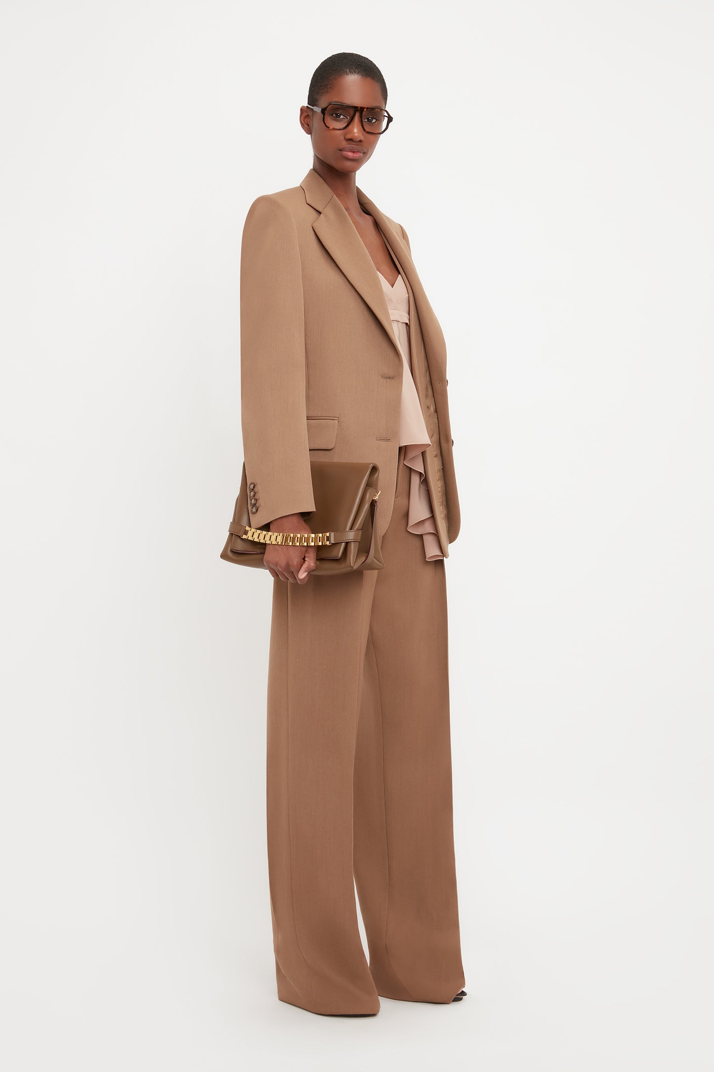 Person standing against a white background, wearing an Asymmetric Double Layer Jacket In Fawn by Victoria Beckham with wide-legged pants, embodying a minimal aesthetic, while holding a brown handbag.