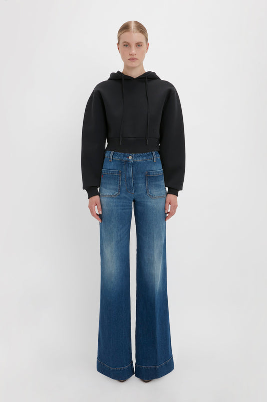 A person stands against a plain background wearing the Cropped Neoprene Hoodie In Black by Victoria Beckham with high-waisted blue wide-leg jeans with front pockets. They have their hair tied back.