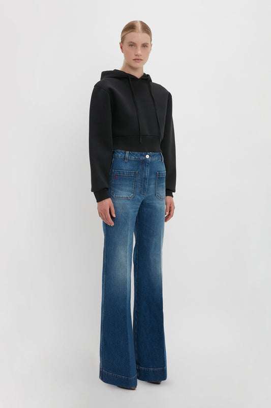 A person stands against a plain background, wearing a Victoria Beckham Cropped Neoprene Hoodie In Black and blue high-waisted, wide-leg jeans.
