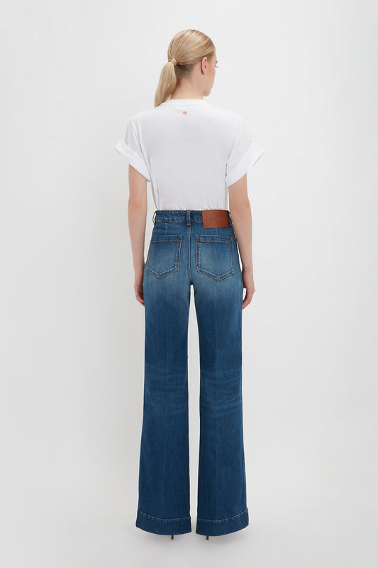 Woman standing with her back to the camera, wearing a white oversized Victoria Beckham Asymmetric Relaxed Fit T-shirt and blue wide-leg jeans with a brown belt, against a plain background.
