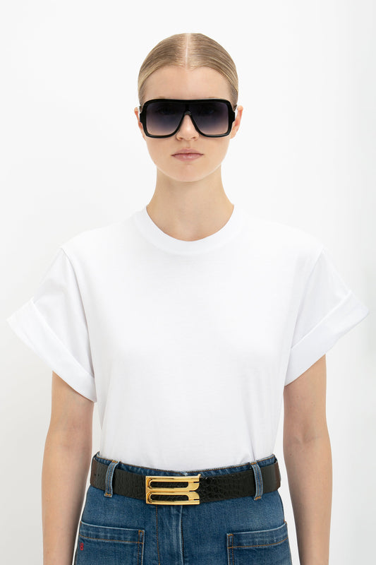 Young woman wearing a Victoria Beckham oversized white t-shirt, blue jeans with a black belt, and large black sunglasses, standing against a white background.