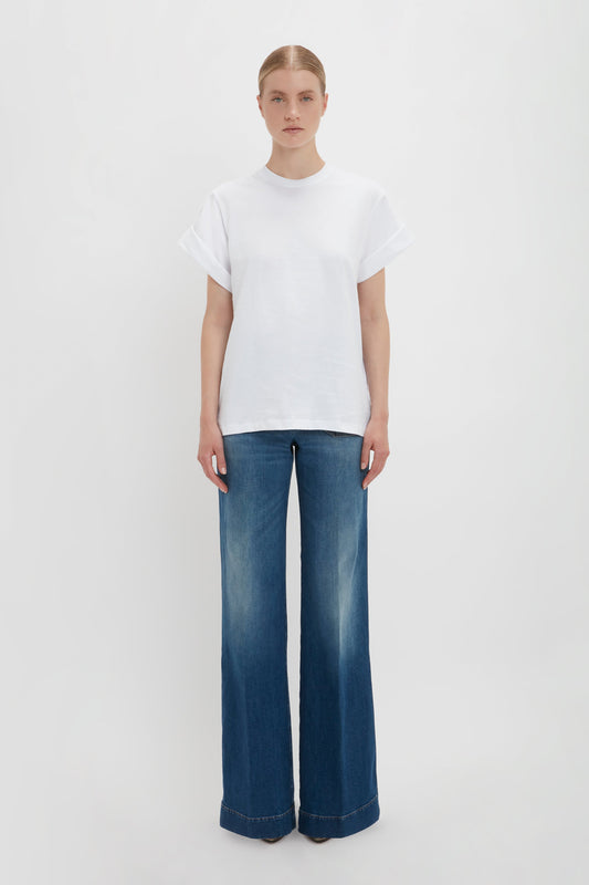 A woman standing against a white background, wearing a white oversized Asymmetric Relaxed Fit T-Shirt by Victoria Beckham and blue Alina jeans in dark vintage wash.