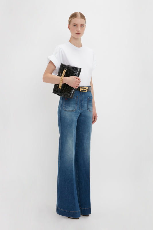 A woman in an oversized Asymmetric Relaxed Fit T-Shirt In White by Victoria Beckham and blue jeans holding a black textured clutch, accessorized with a gold belt, standing against a white background.