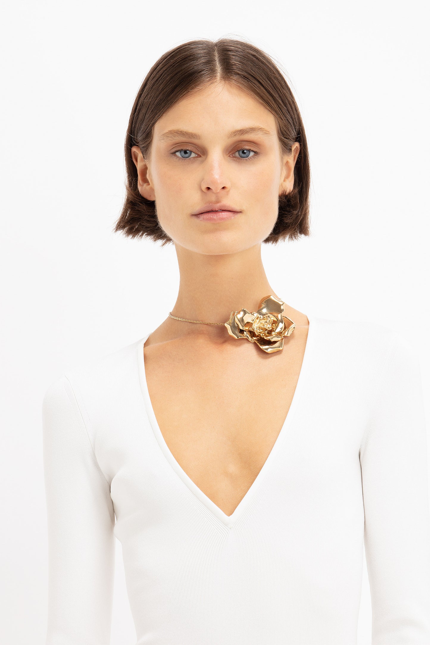 A person with a bob haircut wearing a white V-neck top and a statement accessory: the Exclusive Flower Necklace In Gold by Victoria Beckham, stands against a plain white background.