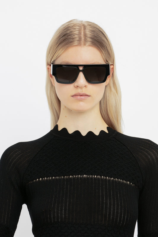 A young woman wearing Victoria Beckham's V Plaque Frame Sunglasses In Black and a black textured top, standing against a white background.