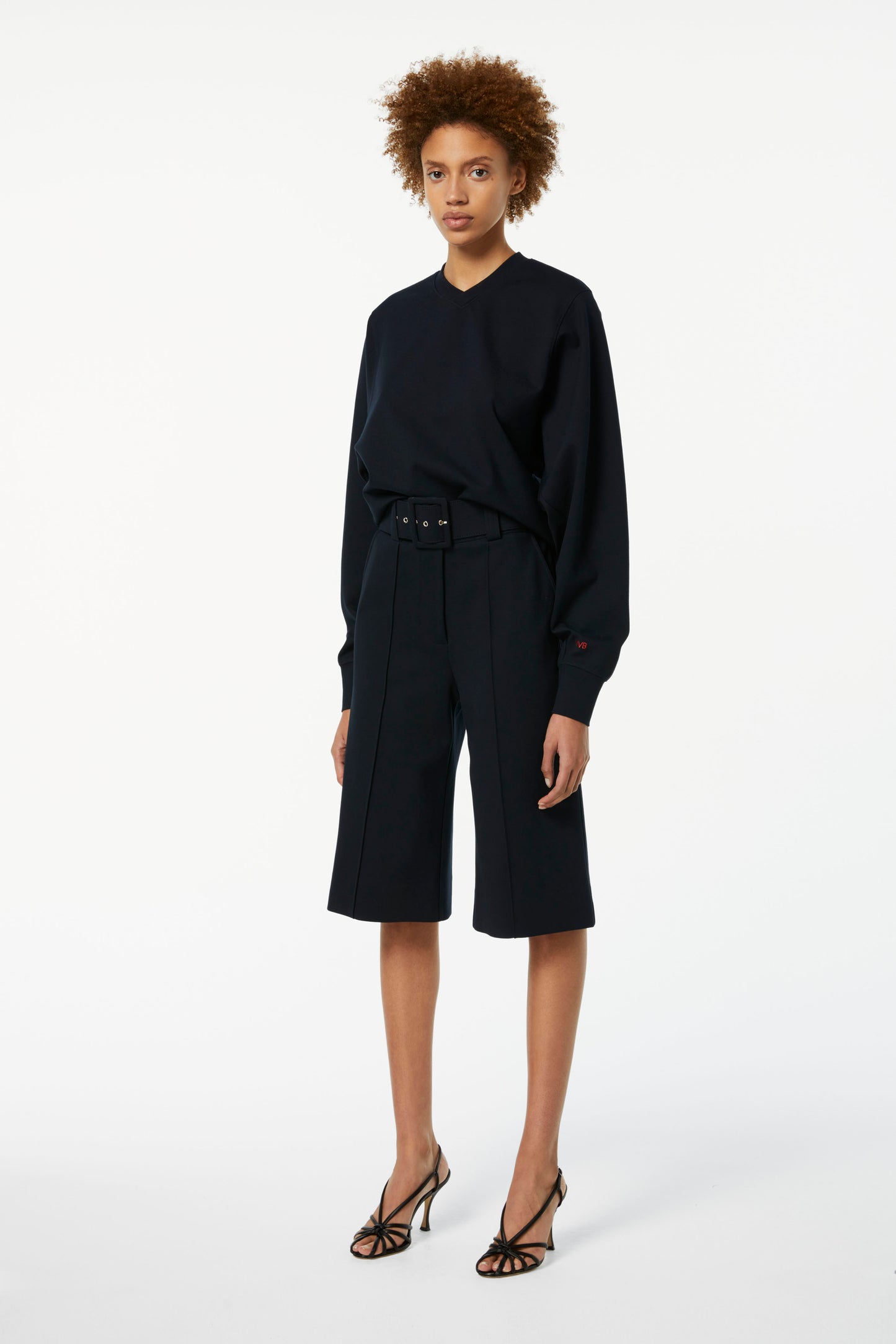 A person with curly hair is wearing a dark long-sleeve top, Victoria Beckham Belted Jersey Culottes in Dark Navy with a flattering fit, and strappy high-heeled sandals, standing against a plain white background.