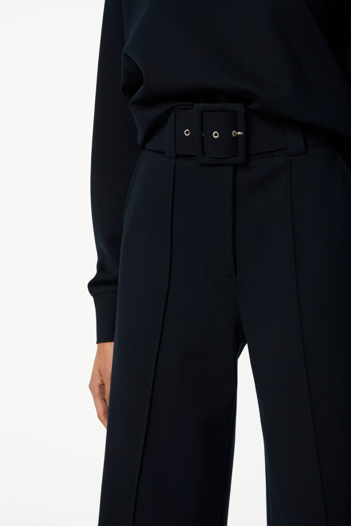 Close-up of a person wearing a black long-sleeved top and flattering Belted Jersey Culottes in Dark Navy by Victoria Beckham with a black belt. Only the midsection and one arm are visible.