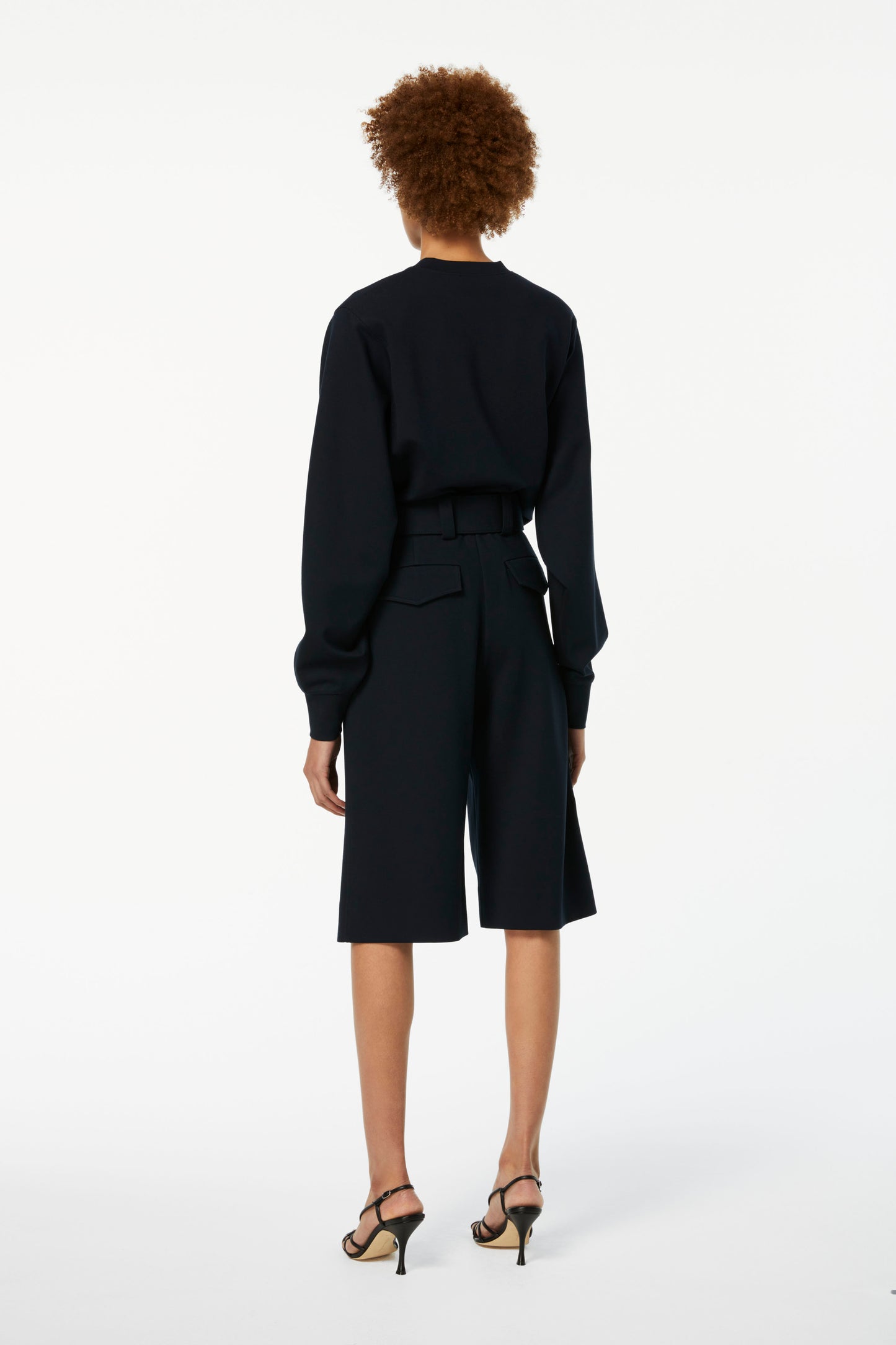 A person with curly hair stands facing away, wearing a black long-sleeved top, Victoria Beckham Belted Jersey Culottes in Dark Navy, and black high-heeled sandals against a plain white background.