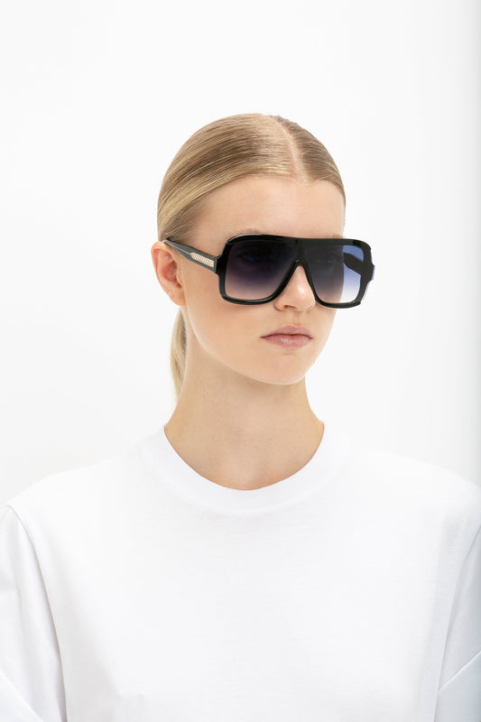 A woman with a pulled-back hairstyle wearing oversized Victoria Beckham black layered mask sunglasses with black lenses and a plain white t-shirt against a white background.
