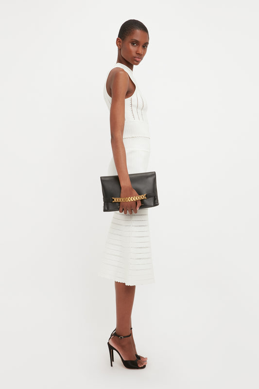 A woman in a white knit dress with a flared midi skirt and black heels carries a Victoria Beckham black chain pouch bag with gold-tone hardware, posing side-on to the camera.
