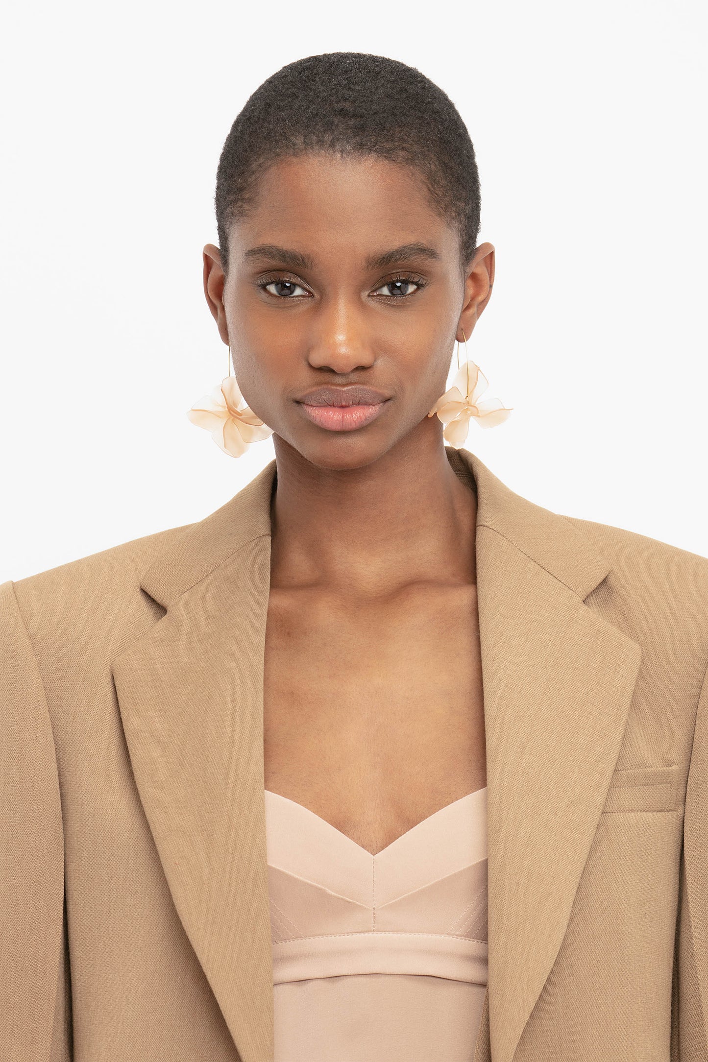 A person wearing an Asymmetric Double Layer Jacket In Fawn by Victoria Beckham over a beige top and large, floral earrings stands against a plain white background, embracing a minimal aesthetic.