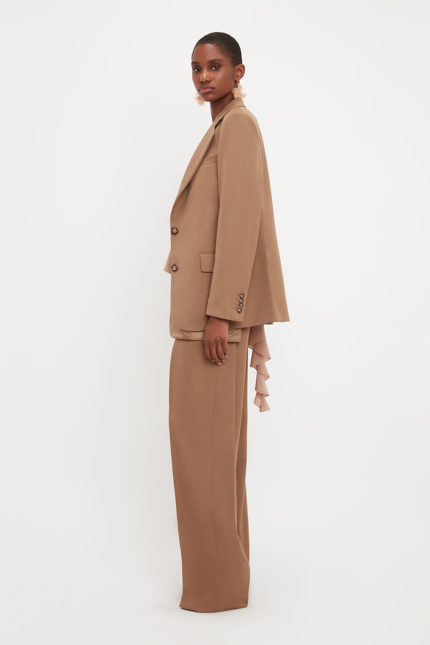 Person in an Asymmetric Double Layer Jacket In Fawn by Victoria Beckham stands against a plain white background, embodying a minimal aesthetic, looking towards the camera.