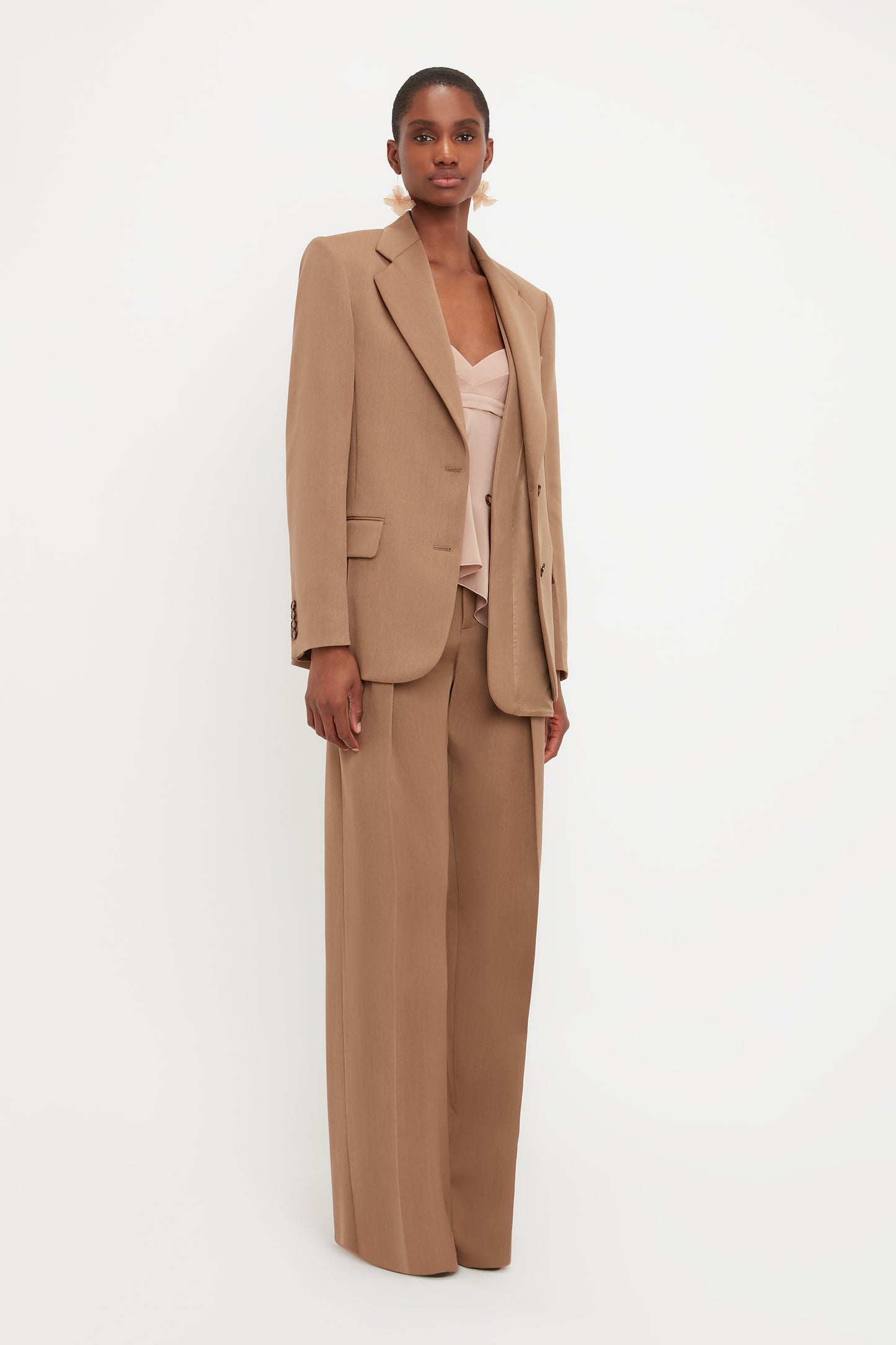 A person stands against a white background, embodying a minimal aesthetic in the Victoria Beckham Asymmetric Double Layer Jacket In Fawn over a light pink top and matching tan wide-leg trousers. They have short hair and are looking directly at the camera.