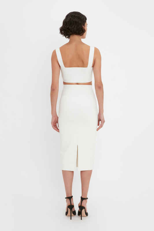 A woman viewed from the back, wearing a white, strapless VB Body Strap Bandeau Top In White and VB Body Fitted Midi Skirt, paired with black high heels, against a plain background.