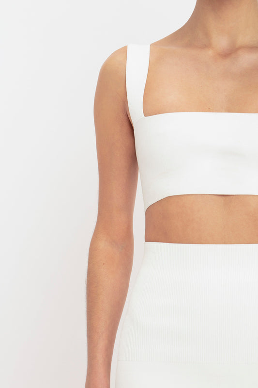 A close-up side view of a person wearing a white crop top with a single shoulder strap and a matching high-waisted Victoria Beckham Body Strap Bandeau Top In White Midi Skirt, against a plain background.