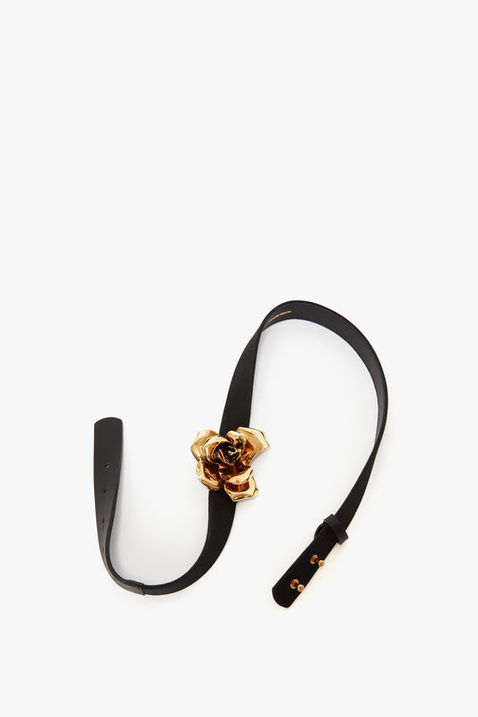A Victoria Beckham exclusive flower belt in black and gold, with a sculptural floral design buckle in gold-plated metal, laid flat on a white background.