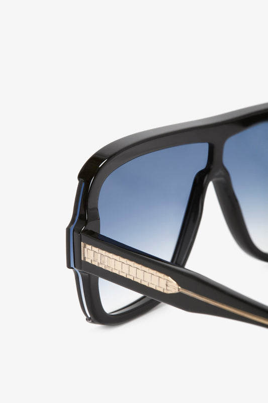 Close-up of Victoria Beckham's Layered Mask Sunglasses In Black Gradient with blue-tinted lenses and a textured metallic detail on the arms, isolated against a white background.