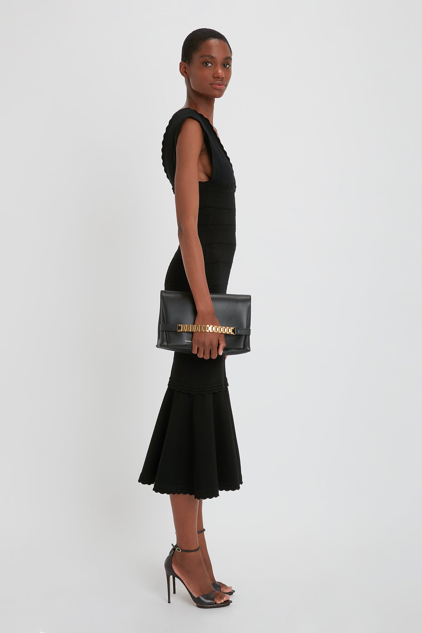 Person standing in profile stance, wearing a sleeveless black dress with a flared hem and black high-heeled shoes, holding a black Chain Pouch Bag with Strap In Black Leather by Victoria Beckham.