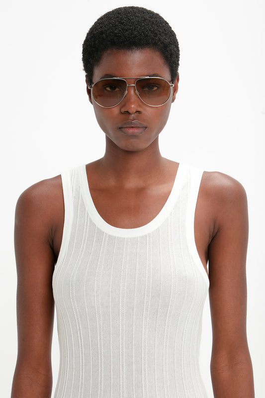 A black woman wearing a white tank top and large Victoria Beckham V Metal Pilot Sunglasses In Silver-Brown, posing against a plain white background.