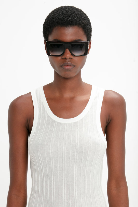 Person with short hair wearing a white sleeveless top and sleek and modern, Victoria Beckham Oversized Frame Sunglasses In Black, looking straight ahead against a plain white background.
