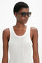 A person wearing a white sleeveless shirt and Victoria Beckham eyewear stands against a plain background, sporting trendy Soft Square Frame Sunglasses In Striped Khaki Havana.