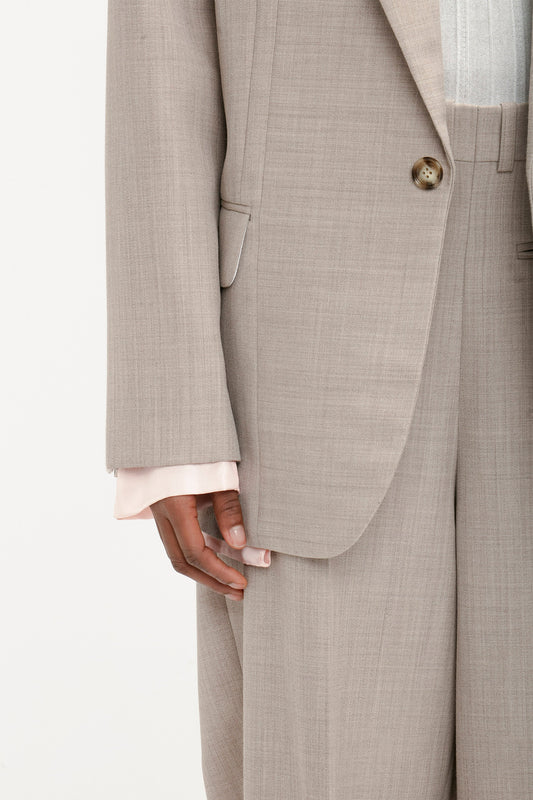 Close-up of a person wearing the Darted Sleeve Tailored Jacket In Sesame from Victoria Beckham with matching trousers. The suit, crafted from pure wool, subtly showcases a light pink shirt sleeve peeking out from under the jacket, highlighting its contemporary detailing.