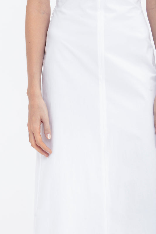 Close-up of a person wearing a white summer dress, showcasing the torso and one arm. The stretch cotton fabric adds a comfortable touch, while the plain white background accentuates its bohemian vibe. The **Cami Fit And Flare Midi In White** by **Victoria Beckham** is elegantly featured in this scene.