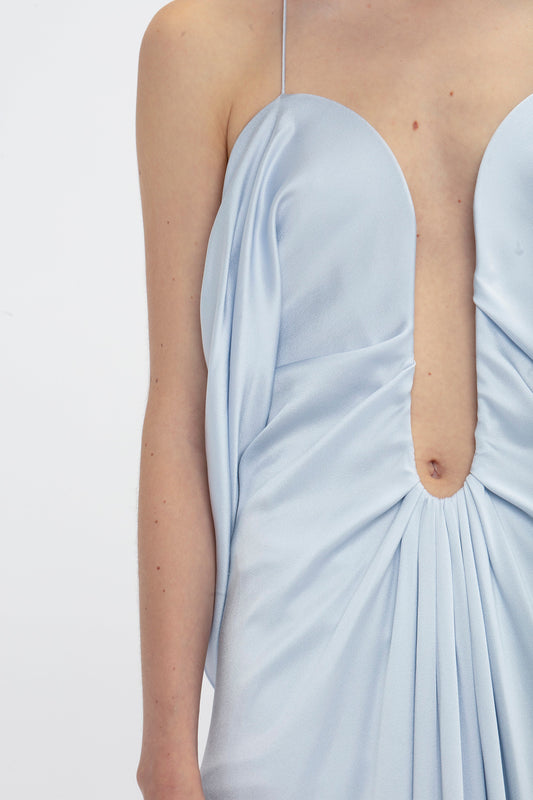 Close-up of a person wearing an elegant light blue Frame Detail Cut-Out Cami Dress In Ice by Victoria Beckham with a deep plunge neckline and draped crepe back satin fabric, showcasing the upper torso and shoulders against a plain white background.