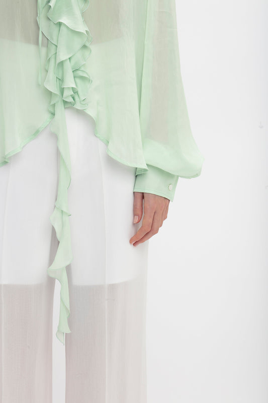A person is shown from chest to mid-thigh wearing a light green Romantic Blouse In Jade with blouson sleeves and white transparent pants from Victoria Beckham. The image focuses on the left hand and hem of the blouse.