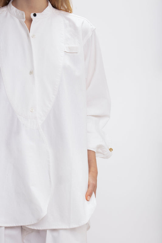 A person wearing a relaxed fit, white button-up Bib-Front Tuxedo Shirt In Washed White by Victoria Beckham with a high grandfather collar and long sleeves stands against a plain background. The shirt has a small pocket at the chest and appears to be tucked in.