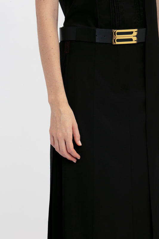 A person wearing a black outfit with a black and gold belt. Only the lower part of their torso and their hand are visible, accentuated by a Victoria Beckham Tailored Floor-Length Skirt In Black that creates a structured silhouette.