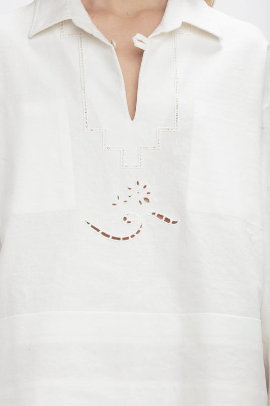 A person is wearing an Oversized Embroidered Tunic In Antique White by Victoria Beckham, featuring an embroidered design with geometric shapes and a swirl in the center. The garment has a V-shaped neckline with a small button closure at the top, reminiscent of Victoria's runway picks.