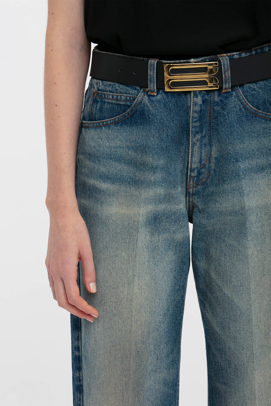 Close-up of a person wearing Victoria Beckham's Relaxed Straight Leg Jean In Antique Indigo Wash with a black belt featuring a golden buckle, focusing on the midsection and right hand.