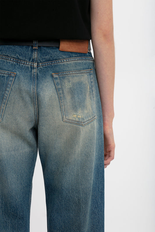 Close-up of a person wearing Victoria Beckham's Relaxed Straight Leg Jean In Antique Indigo Wash, focusing on the back pocket and brown belt against a white background.