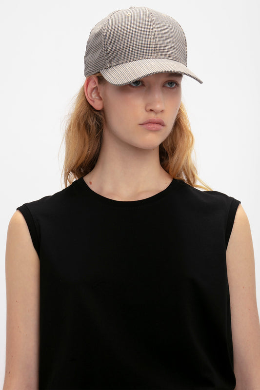 Young woman wearing a black sleeveless top and a gray Victoria Beckham Logo Cap In Dogtooth Check, looking to the side with a neutral expression against a plain background.